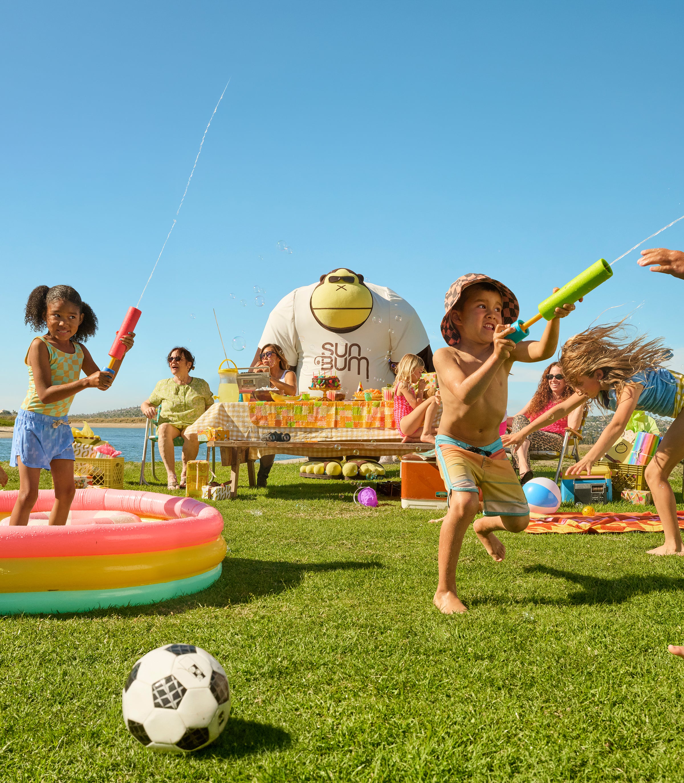 Children playing with water guns and a soccer ball on a grassy lawn with a pool and people in the background.