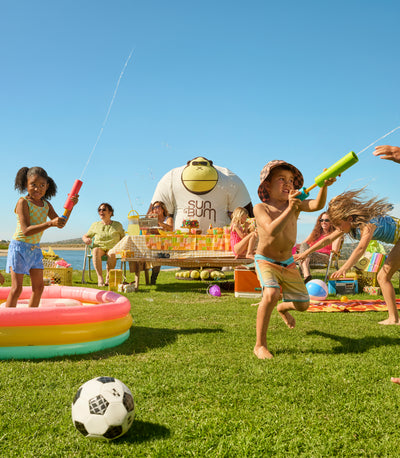 Children playing with water guns and a soccer ball on a grassy lawn with a pool and people in the background.