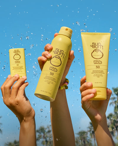 Three bottles of Sun Bum Kids sunscreen being held up against a blue sky.