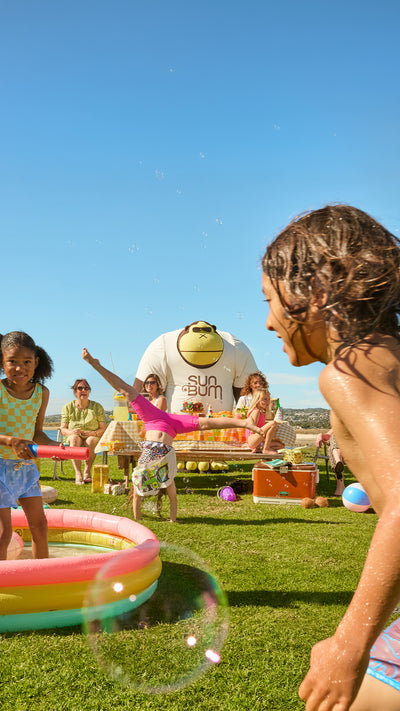 Children playing with bubbles in a grassy area with a pool and inflatable slide in the background.