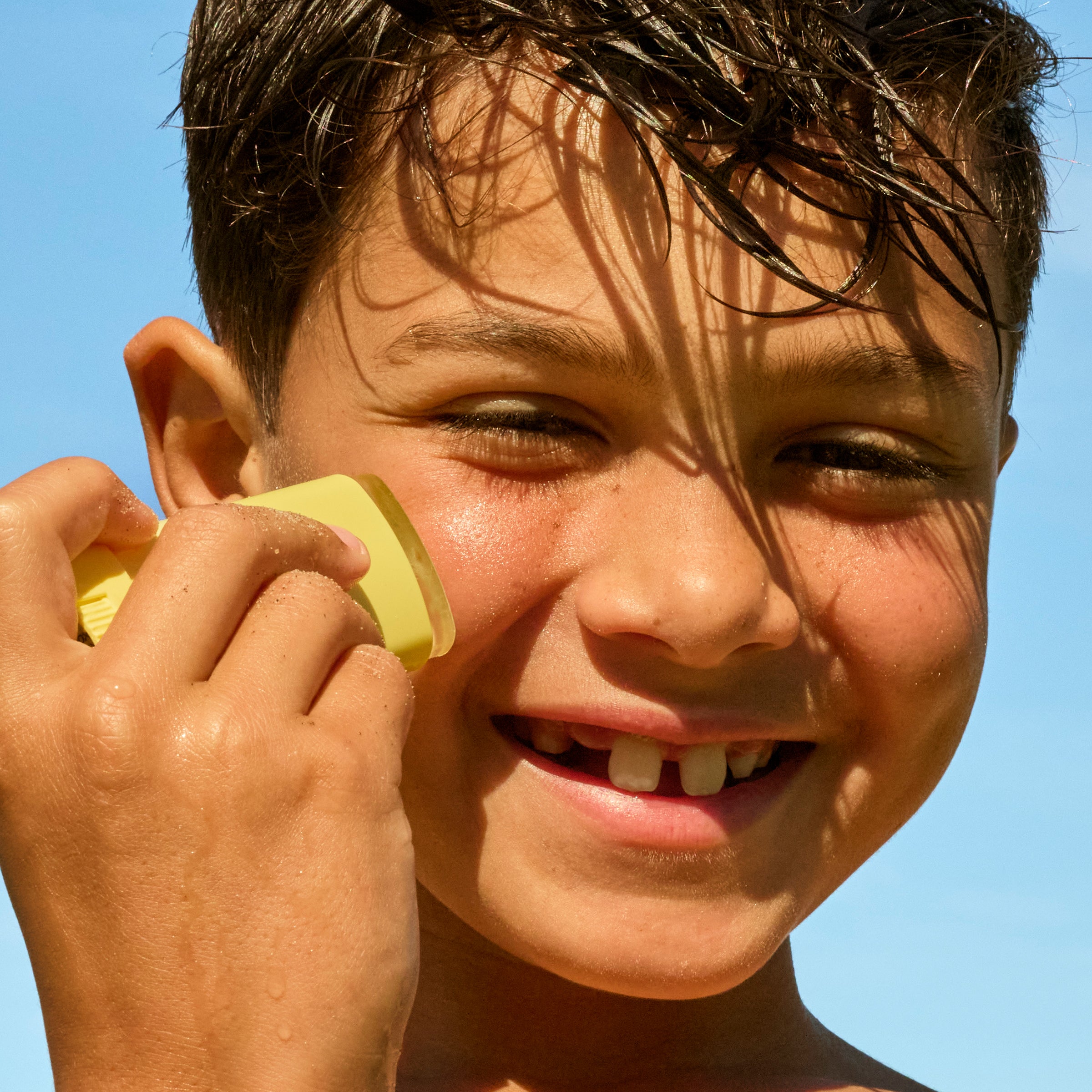 Little boy putting on sunscreen face stick on his face on a blue sky background. 