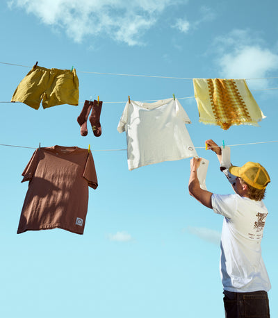 A person in a white t-shirt and yellow cap hangs a white t-shirt on a clothesline outdoors against a bright blue sky, with other clothes including yellow shorts, a brown t-shirt, and socks also hanging.