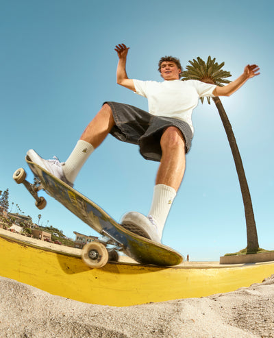 A skateboarder in a white t-shirt and dark shorts performs a trick on a yellow ramp under a clear blue sky with a palm tree in the background.