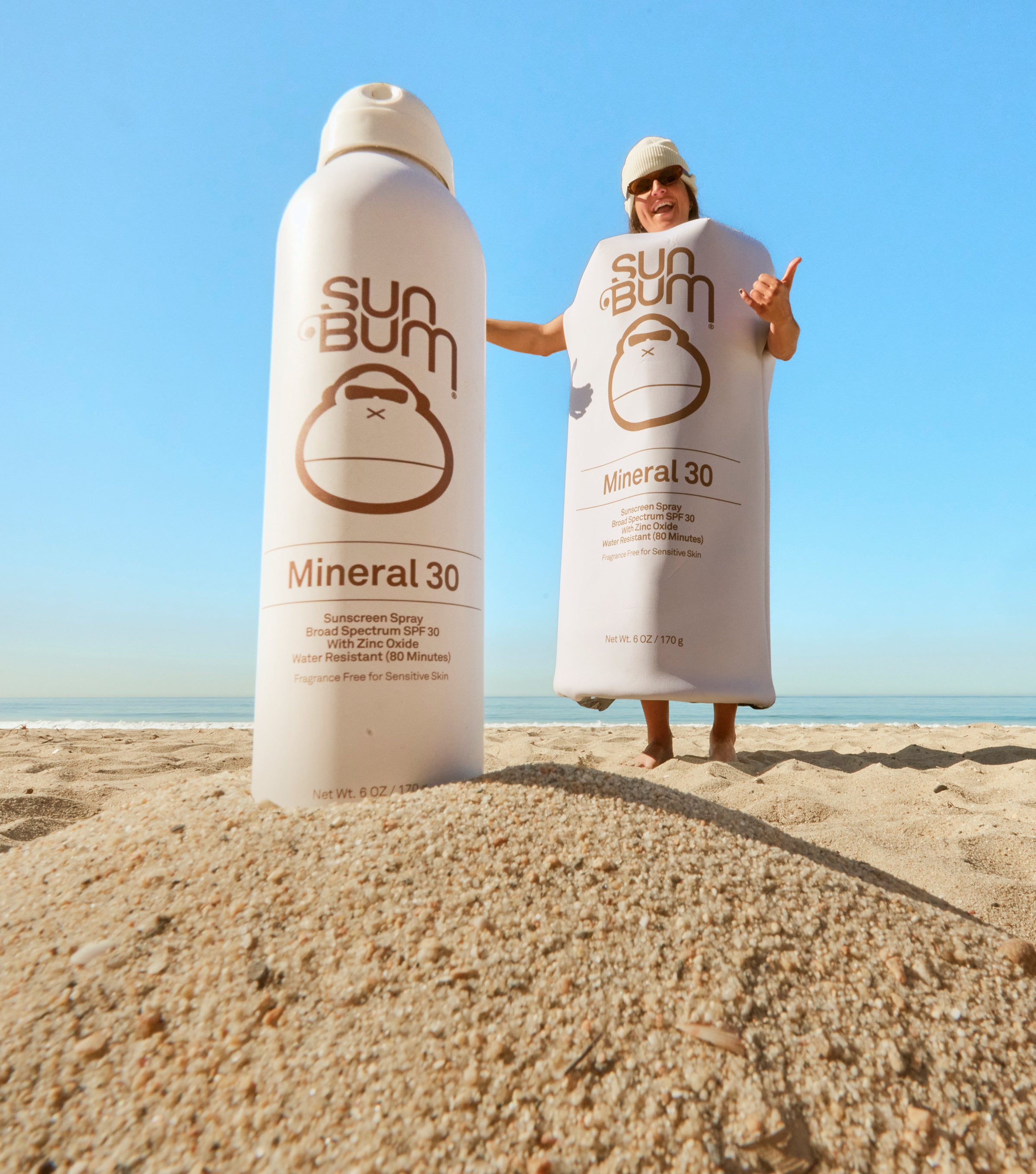 Person wrapped in a Sun Bum towel on a beach with a large bottle of Sun Bum Mineral 30 sunscreen.