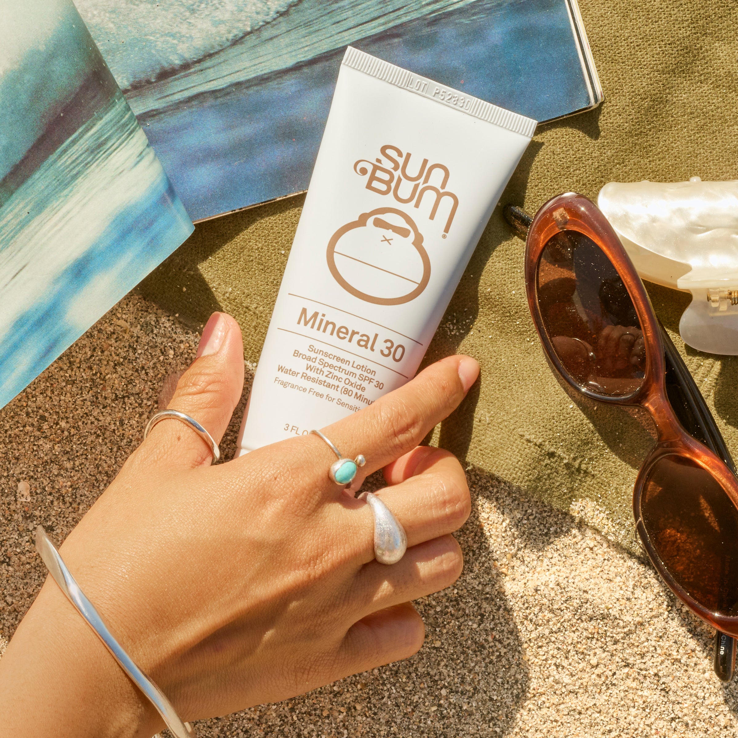 Hand holding a Sun Bum Mineral 30 sunscreen tube on sand with sunglasses and beach towel in the background