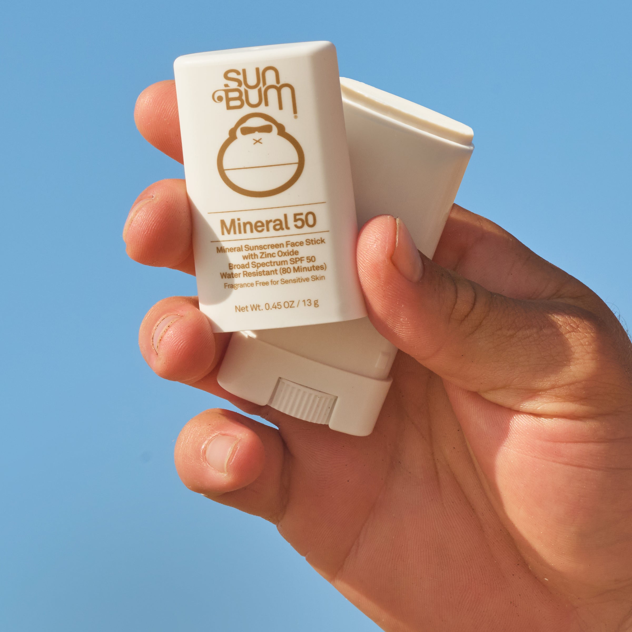 Hand holding a Sun Bum Mineral 50 sunscreen stick against a clear blue sky