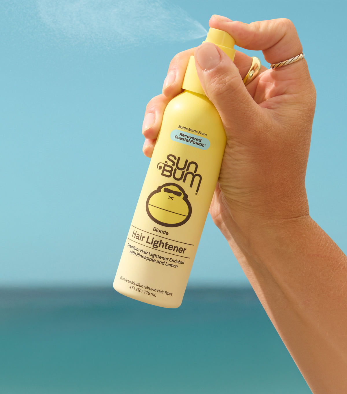 A hand holding a yellow Sun Bum Reef Hair Lightener spray bottle, with the nozzle spraying a fine mist against a bright blue sky background.