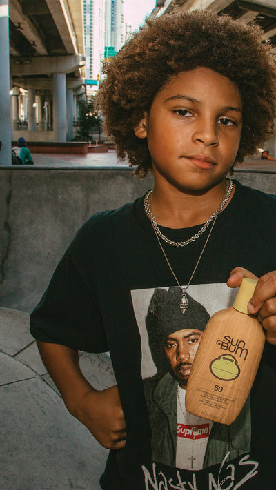 A young boy with an afro hairstyle stands outdoors holding a bottle of Sumo Sunscreen. He is wearing a black graphic t-shirt with a print of Nas and a gold chain necklace.
