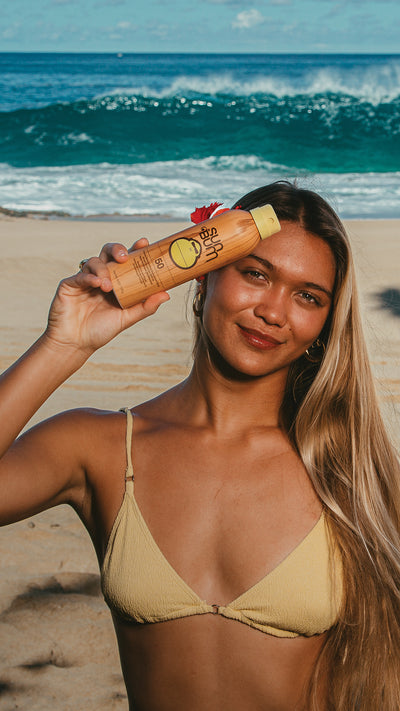 A woman on a sandy beach holds a bottle of sunscreen in front of her face, with ocean waves and blue sky in the background. She is wearing a beige bikini top.