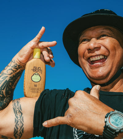 A smiling man outdoors under a bright blue sky holds up a bottle of Sun Bum sunscreen. He is wearing a black wide-brim hat, a black t-shirt, and a digital watch, and making a shaka sign with his hand.