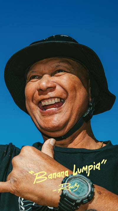 Person wearing a black hat and black shirt with 'Banana Lumpia' quote, giving a shaka up against a blue sky.