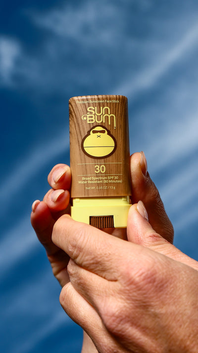Hand holding a Sun Bum sunscreen bottle against a blue sky background