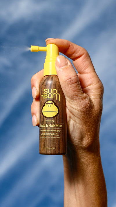 Hand holding a Sun Bum hair mist bottle against a blue background