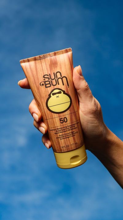 Hand holding a Sun Bum sunscreen tube against a blue sky background