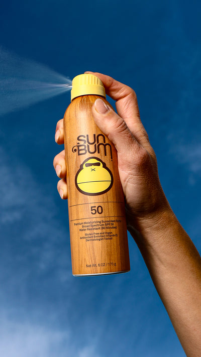 Hand holding a Sun Bum sunscreen spray bottle against a blue sky background