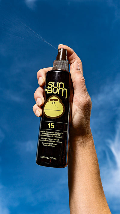 Hand holding a Sun Bum sunscreen spray bottle against a blue sky background