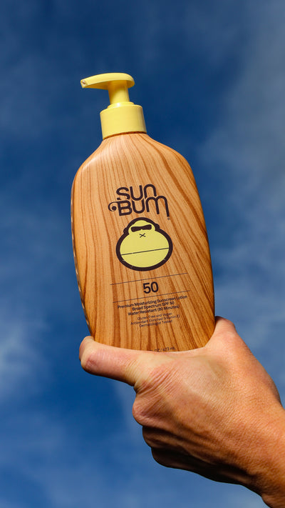 Hand holding a Sun Bum sunscreen bottle against a blue sky background