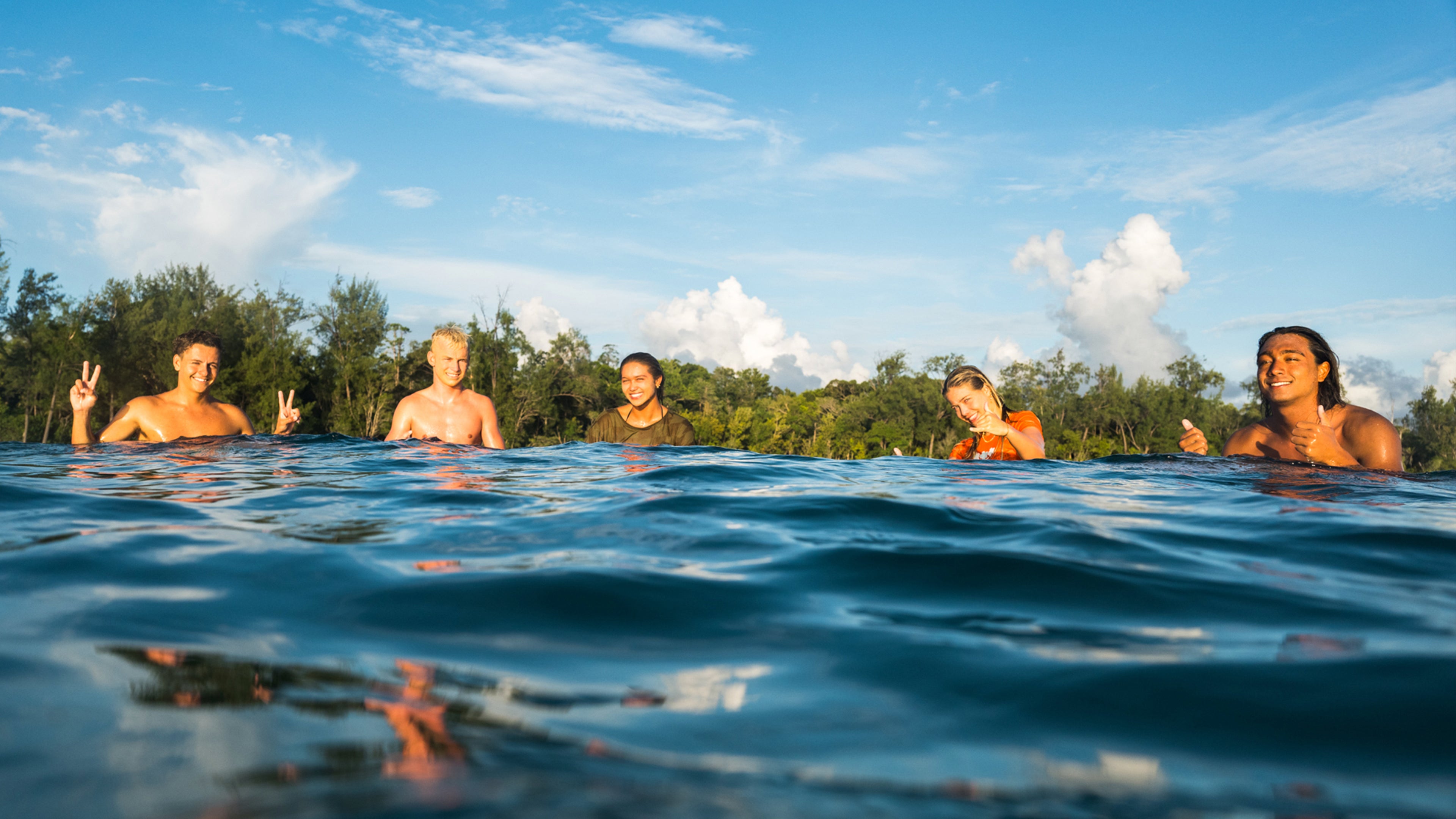 People sitting in water with a scenic background of trees and blue sky.
