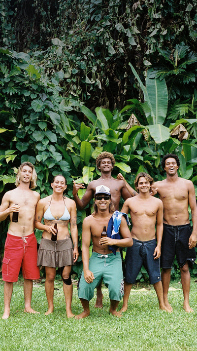 Six men standing together outdoors on grass in front of dense tropical green foliage, wearing swim trunks and a bikini top. One man in the center wears teal swim trunks and a white cap, holding a towel.