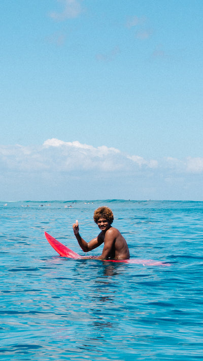A man sitting in turquoise ocean water holding a bright pink surfboard and giving a thumbs-up, with a blue sky and distant clouds behind him.