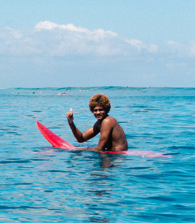 A shirtless man with curly hair sits in turquoise ocean water on a red surfboard, holding up a small white object, with the sea and cloudy sky behind him.