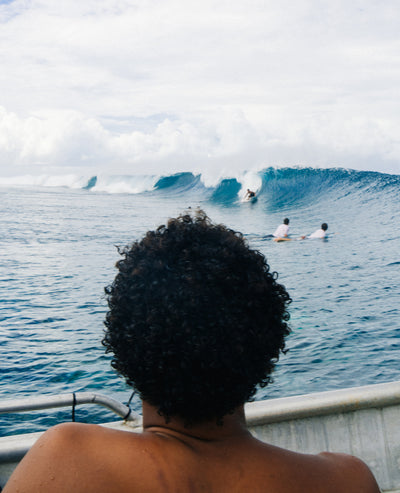 A person with curly dark hair seen from behind, sitting on a boat and watching surfers ride a blue ocean wave under a cloudy sky.