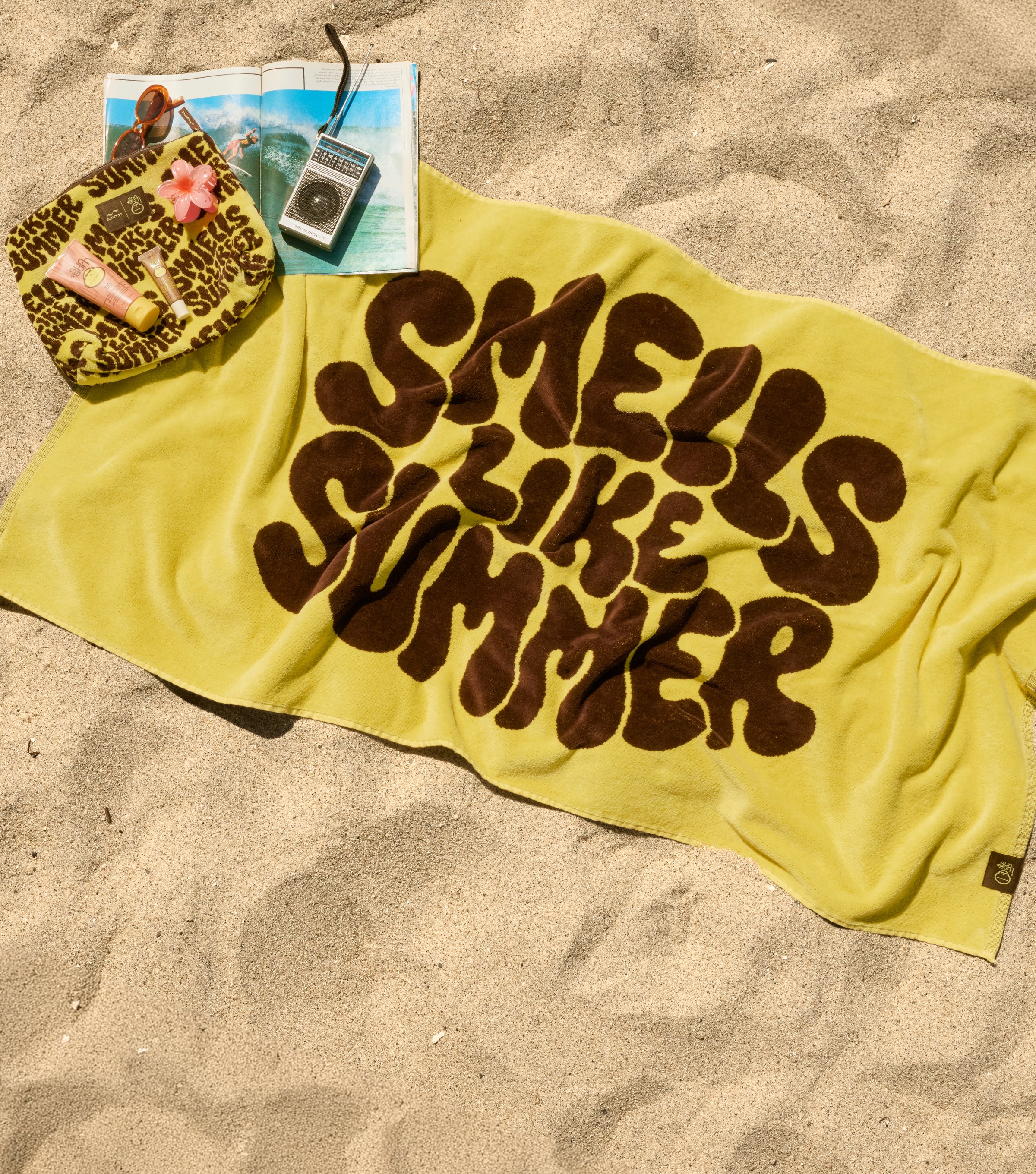 Yellow towel with 'Smells Like Summer' text on sand, next to a leopard print bag and vintage radio.