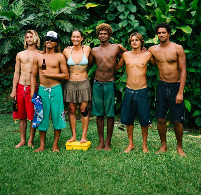 Six friends in swimwear standing together on grass in front of lush tropical foliage