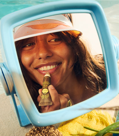 Person holding a lip balm in front of a mirror with a beach background