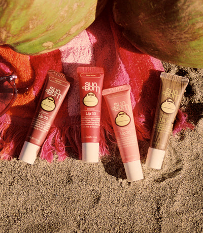 Four tubes of Sun Bum lip balm on sand with a red towel and green leaves in the background