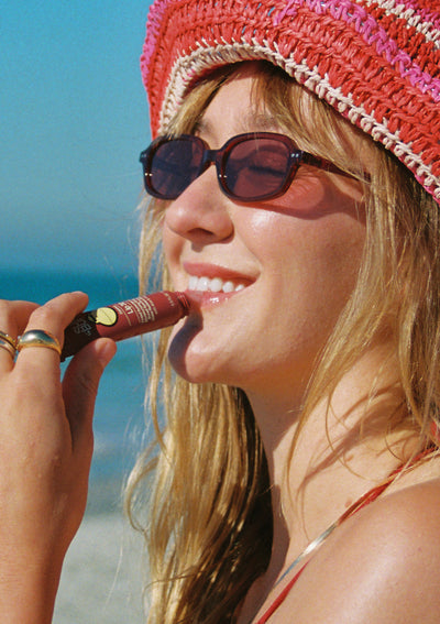 Woman on a beach wearing sunglasses and a pink hat, holding a lip balm.