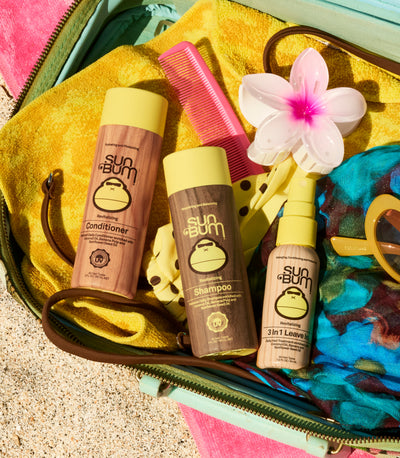 Three Sun Bum hair care products arranged in an open turquoise suitcase with a yellow towel, pink comb, white flower, sunglasses, and colorful accessories. The products are conditioner, shampoo, and a 3-in-1 leave-in.