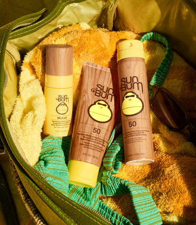 Three Sun Bum sunscreen products arranged inside an olive green bag with yellow towels and sunglasses around them. The products include a small bottle, a squeeze tube, and a larger bottle, all with wood-grain style labels and yellow caps.
