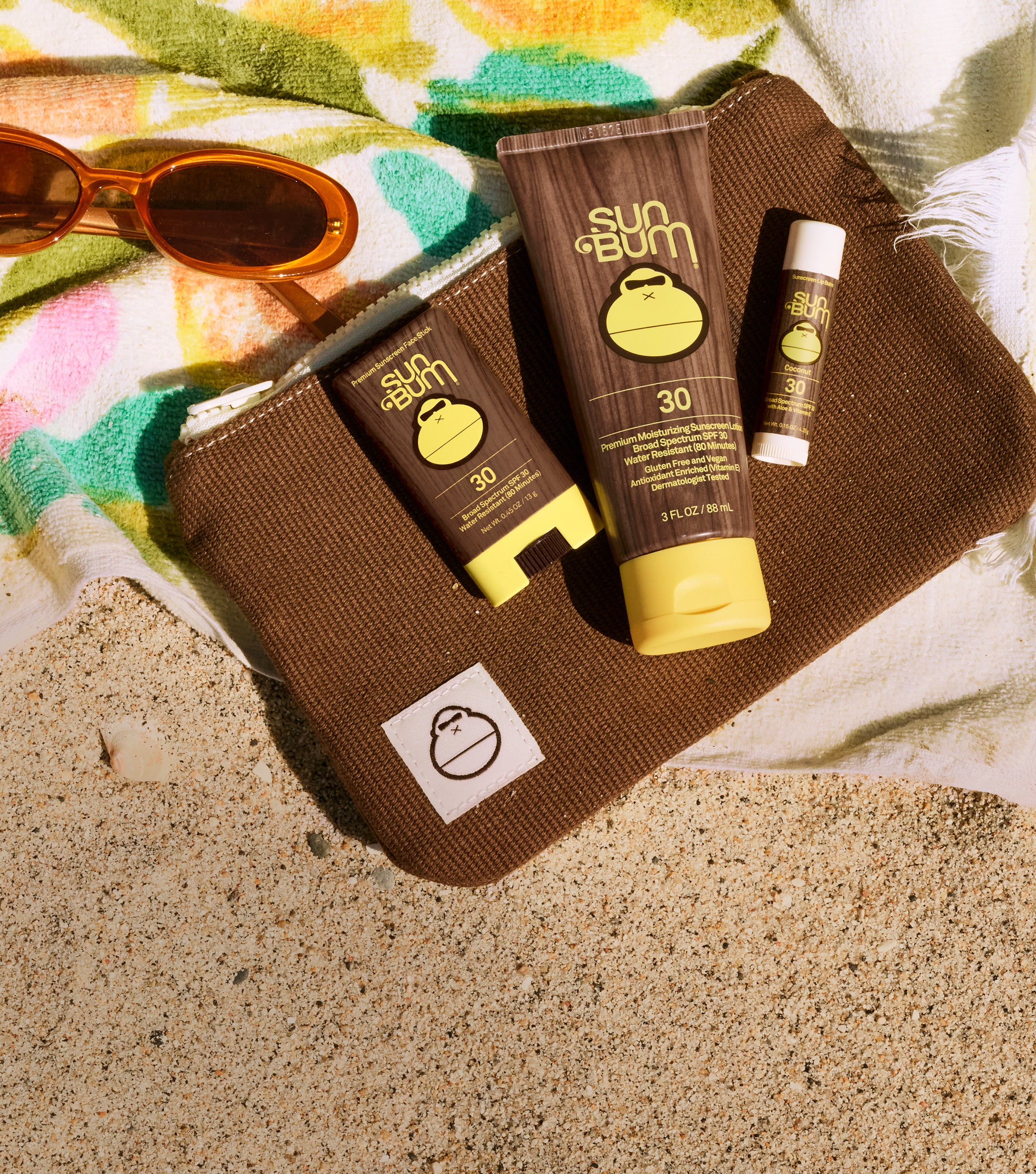 Sun protection products arranged on a brown zip pouch on a beach towel and sand: a large Sun Bum sunscreen tube, a smaller Sun Bum sunscreen tube, a lip balm tube, and tortoiseshell sunglasses.
