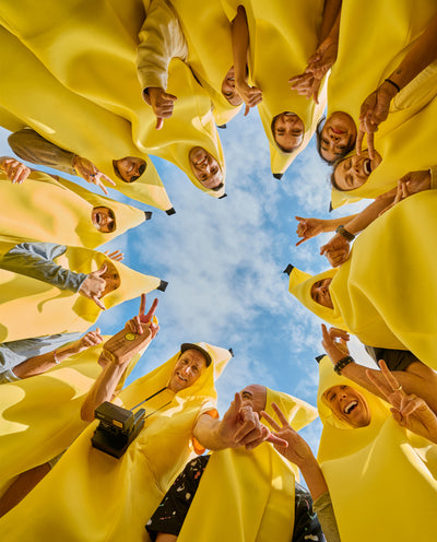 Group of people in banana costumes smiling and posing in a circle under blue sky - Sun Collection - Sun Bum