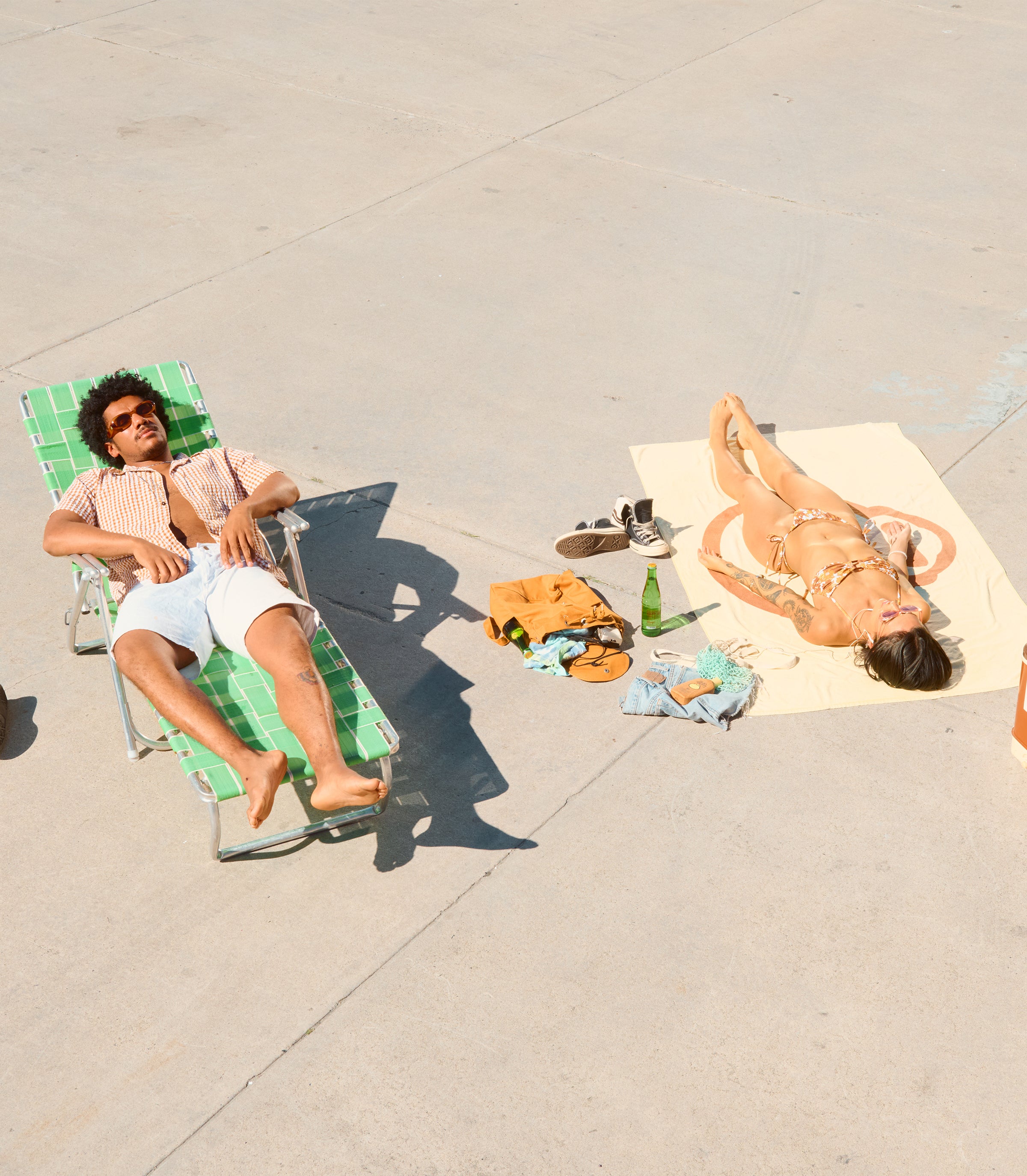 Two friends sunbathing on a sunny pavement with laid-back beach gear, featuring the SB Bestsellers Collection – Sun Bum.