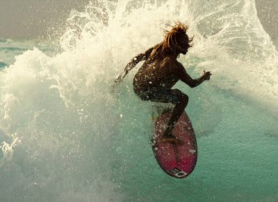 Surfer catching air off a wave at golden hour, showcasing the energy of the Discover Ambassador Collection - Sun Bum.