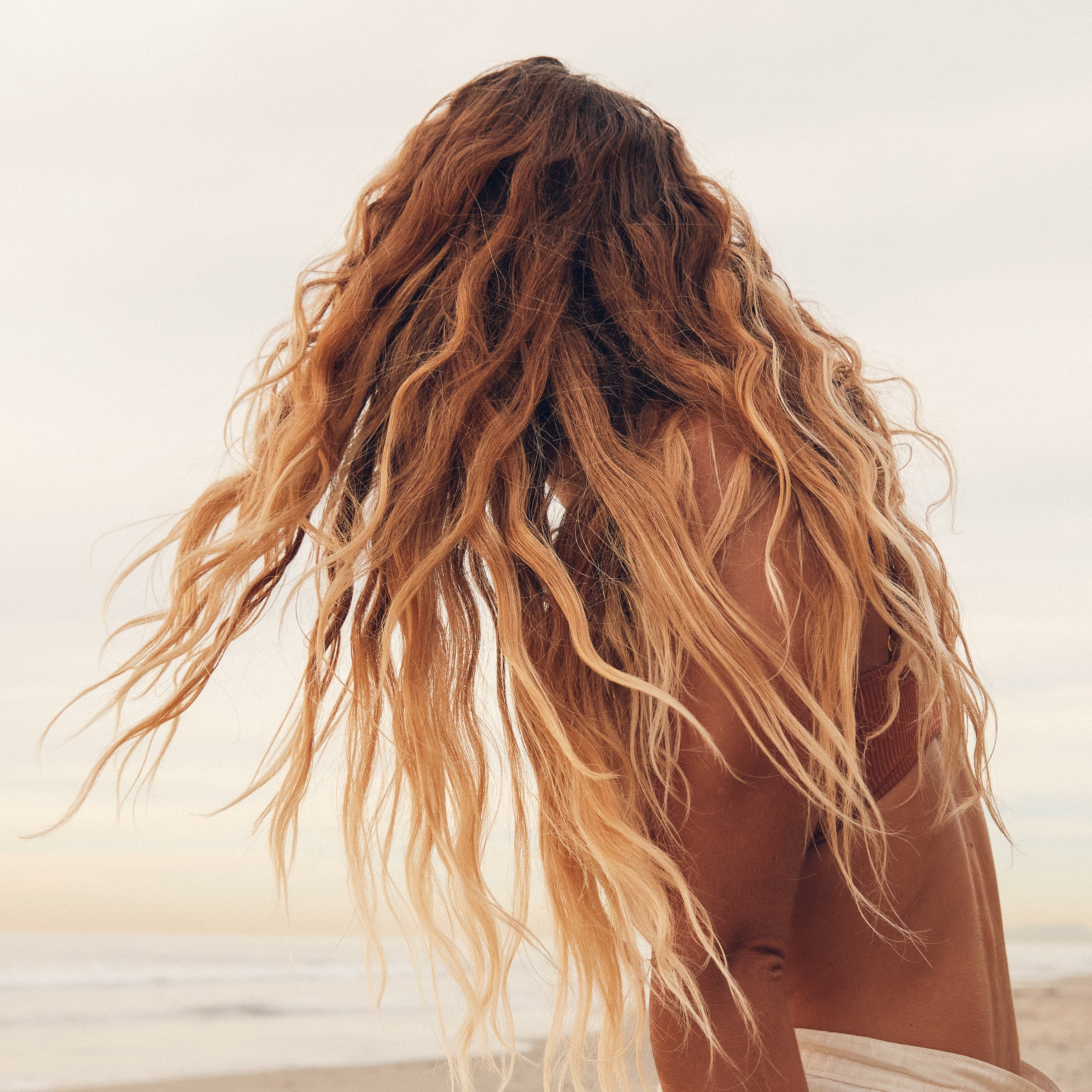 Beachy waves flow freely as a woman tosses her hair near the shoreline – Curls Collection - Sun Bum.
