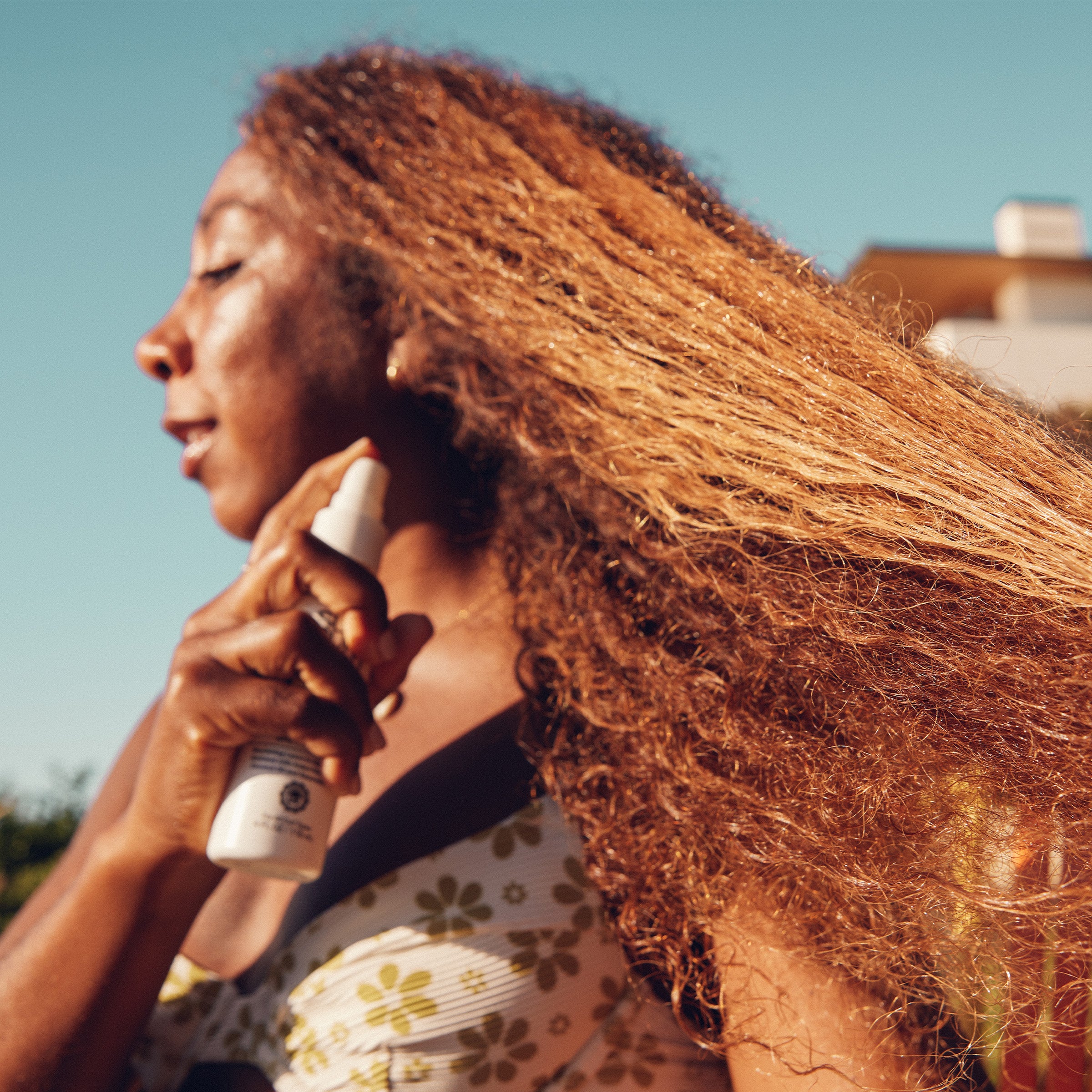 Woman spraying product into long curly hair under the sun for beach-ready protection – Curls Collection - Sun Bum.