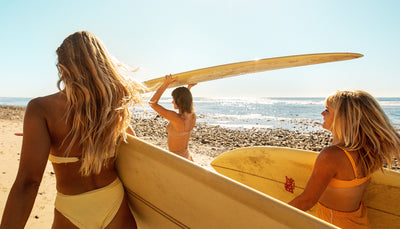 Women carrying surfboards toward the ocean under the sun, showing off healthy beach hair – Hair Collection - Sun Bum.