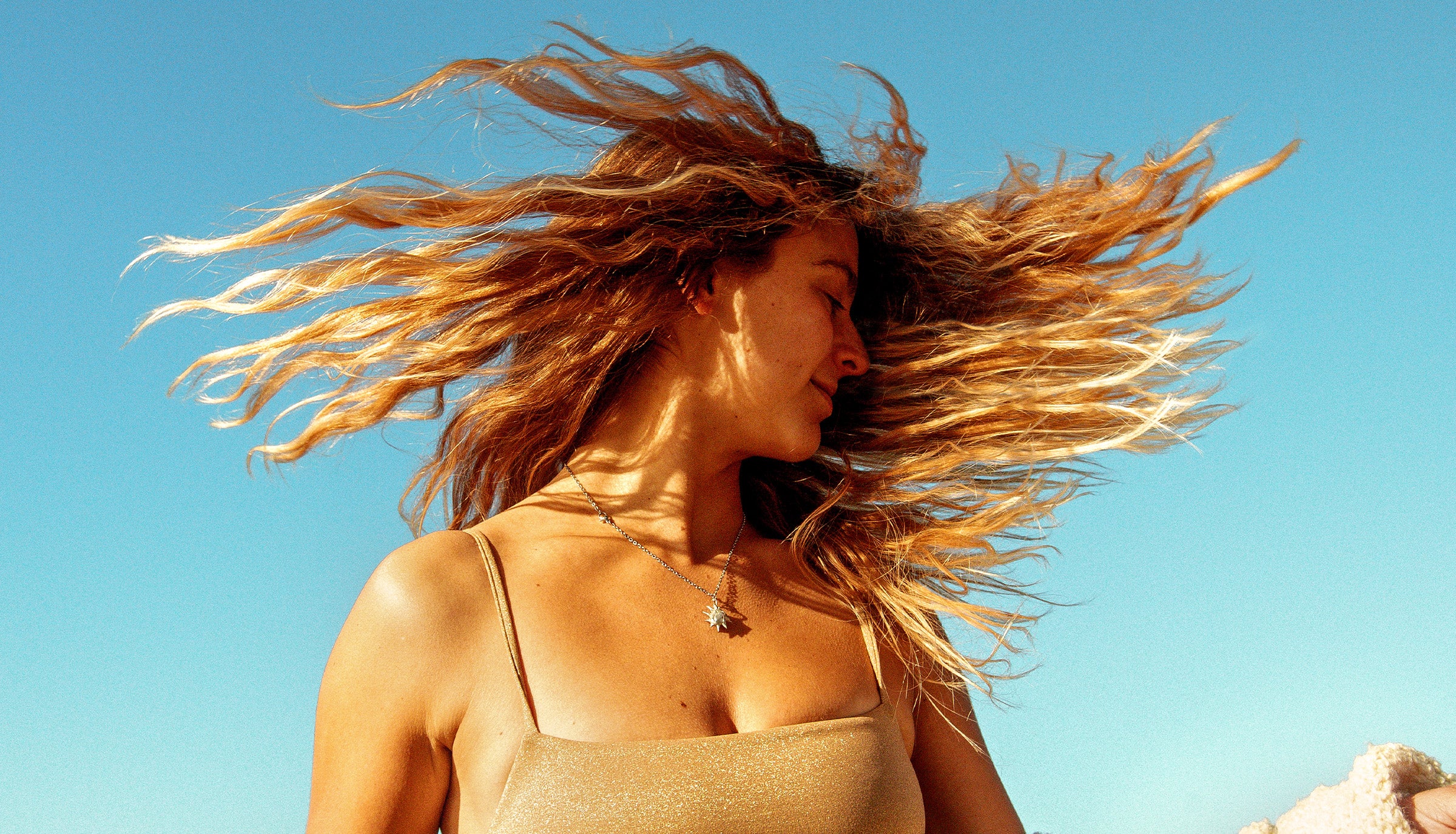 Woman flipping sun-kissed hair under blue sky, showcasing beachy texture and shine – Hair Collection - Sun Bum.