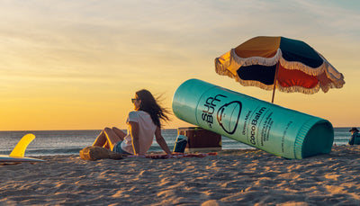 Woman relaxing on the beach at sunset next to a giant CocoBalm lip balm prop – CocoBalm Collection - Sun Bum.