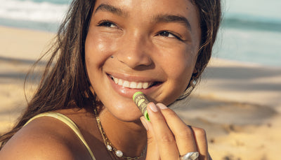 Smiling woman applies SPF 30 lip balm at the beach for sun-safe, hydrated lips – OG Lip Balm Collection - Sun Bum.
