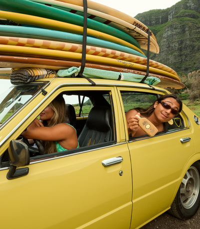 Friends on a surf trip with boards stacked on a yellow car, holding sunscreen lotion from the Lotions Collection – Sun Bum.