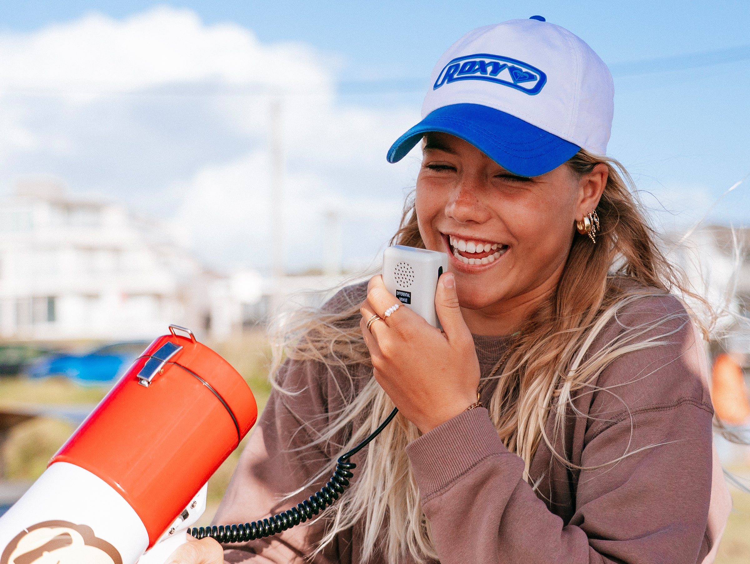 Person wearing a cap with a brand logo, holding a megaphone and a microphone outdoors.