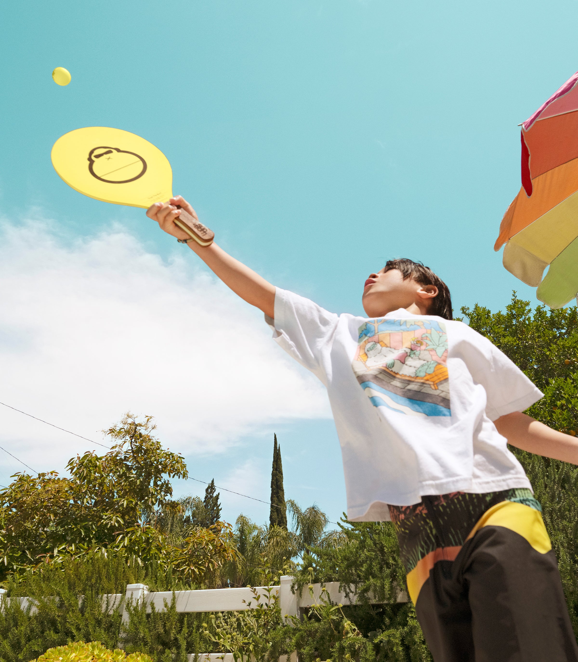 Kid playing paddle ball under a sunny sky in a colorful backyard scene from the Outside Is In Collection – Sun Bum.