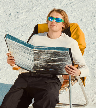 Person sunbathing in snow with reflective visor and sunglasses, promoting SEP Snow Collection - Sun Bum.