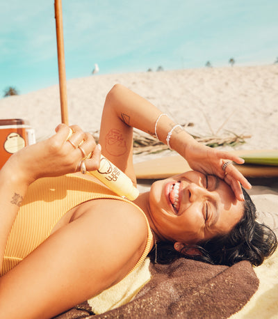 Woman laughing on the beach while applying SPF lotion, featuring the Summer Essentials Collection – Sun Bum.