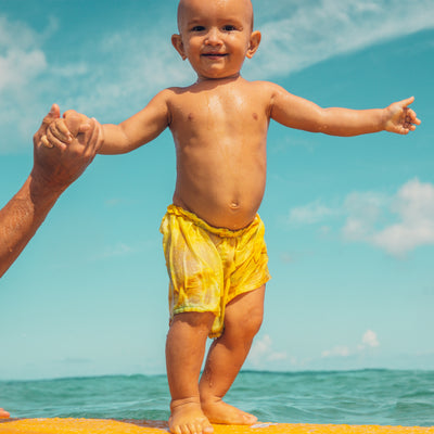 Smiling baby in yellow shorts standing on a surfboard with support at the beach – Baby Bum Collection - Sun Bum.