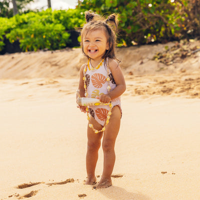 Smiling toddler on the beach holding Baby Bum sunscreen and wearing a seashell necklace – Baby Bum Collection - Sun Bum.